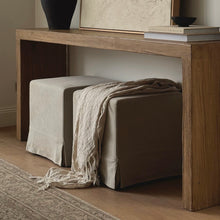 Load image into Gallery viewer, A rustic wooden console table in a warm brown finish, styled with a beige slipcovered storage ottoman and a cream fringed throw blanket, set in a cozy room with a patterned rug.
